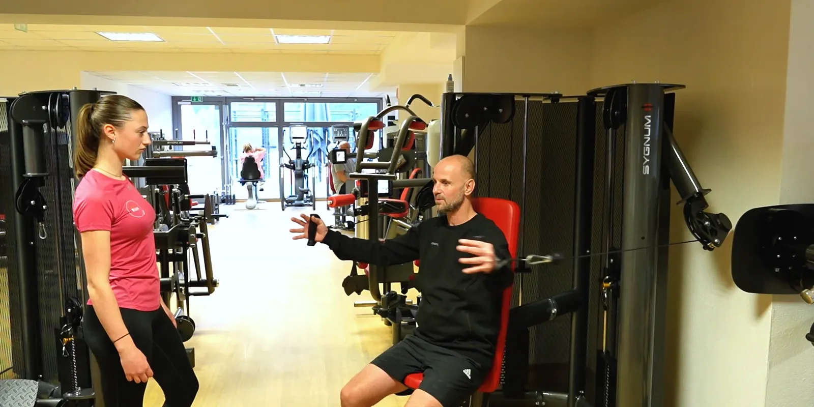 Trainer erklärt einem Mitglied die Kraftgeräte